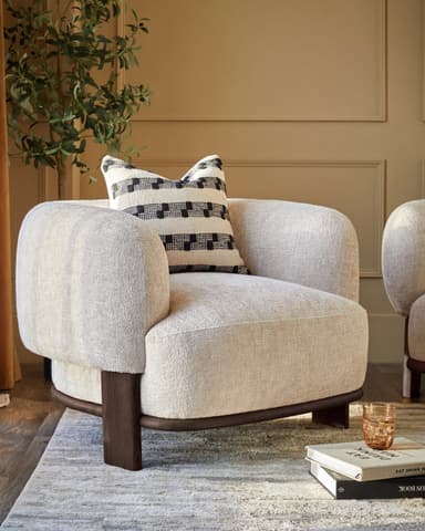 An upholstered armchair featuring a rounded, barrel-like silhouette with integrated armrests and a dark wood plinth base. The chair is covered in a light, textured fabric and is accompanied by a patterned throw pillow. — 3D model