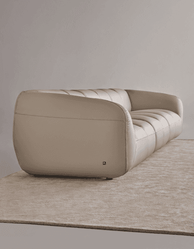A contemporary four-seat sofa with a low profile and rounded arms. The sofa is upholstered in a light beige or cream-colored material, likely leather or a high-quality faux leather, and features prominent stitching that delineates the seat and back cushions. The design is modular, with two distinct seating sections joined together. It is supported by small, dark, cylindrical feet. — 3D model