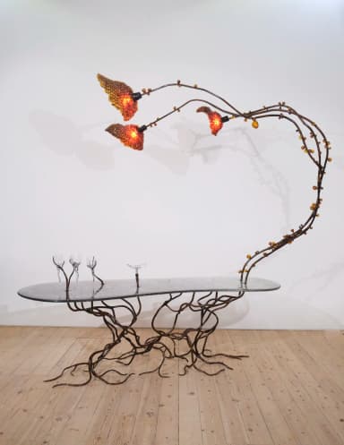 A highly sculptural coffee table featuring an elongated, oval-shaped glass top and a patinated metal base sculpted to resemble intertwined roots. An integrated light fixture, designed as an arching branch with three organic, textured shades and amber glass beads, extends from the base over the tabletop. — 3D model