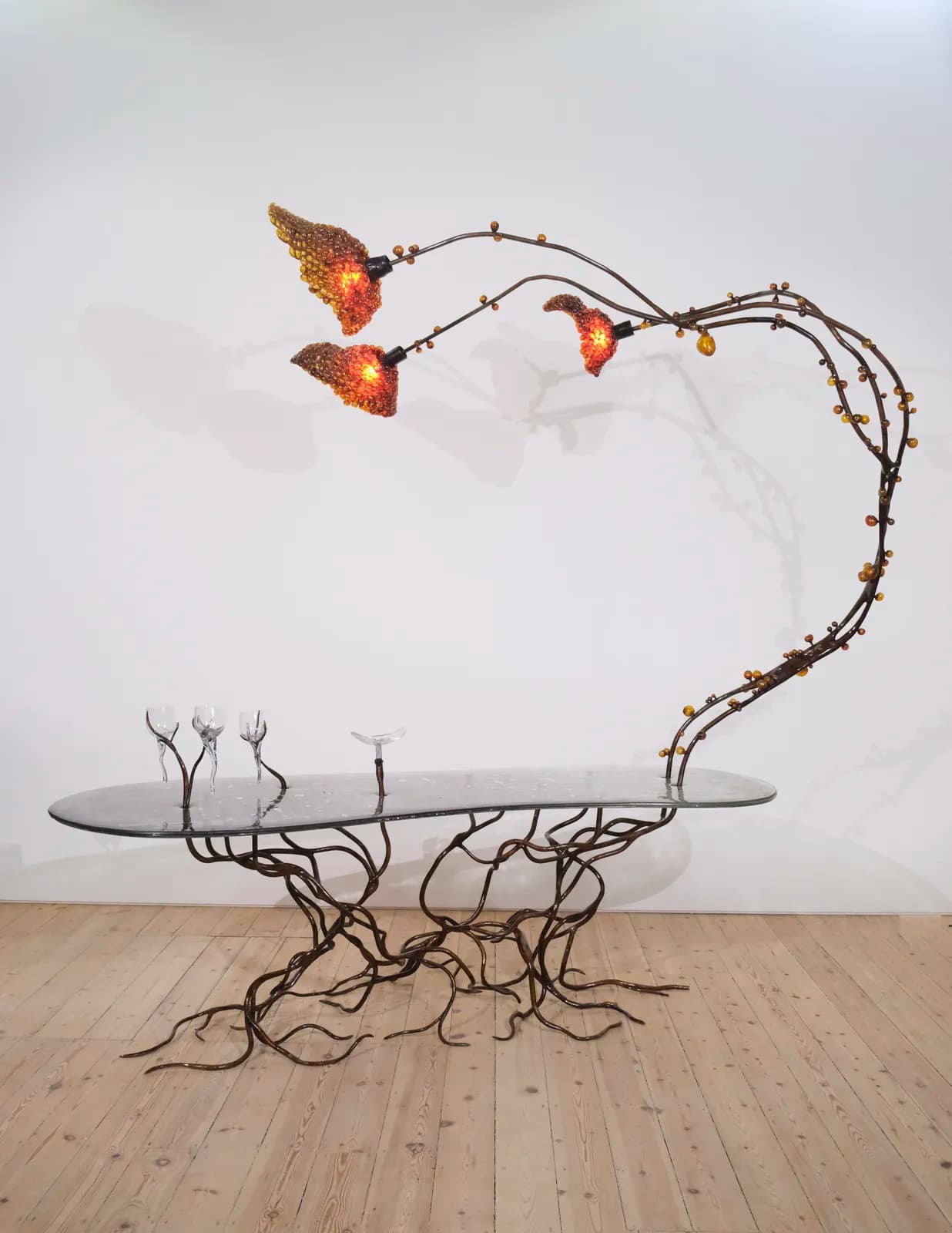 A highly sculptural coffee table featuring an elongated, oval-shaped glass top and a patinated metal base sculpted to resemble intertwined roots. An integrated light fixture, designed as an arching branch with three organic, textured shades and amber glass beads, extends from the base over the tabletop. — 3D model