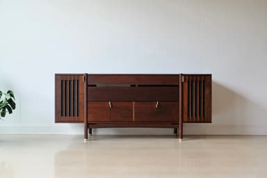 A long, low, dark wooden cabinet with a distinct mid-century modern design. It features slatted sections on the curved ends, a central open shelf, and multiple drawers and cabinet doors below, all adorned with small brass pull handles. The cabinet is supported by angled, tapered legs with brass ferrules at the bottom. — 3D model