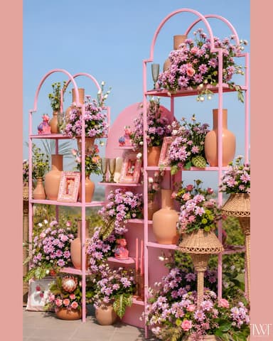 A decorative, freestanding shelving unit constructed from pink-painted metal, featuring a multi-tiered design with elegant arched tops. The unit is styled with numerous floral arrangements in terracotta vases, picture frames, and small decorative objects. — 3D model