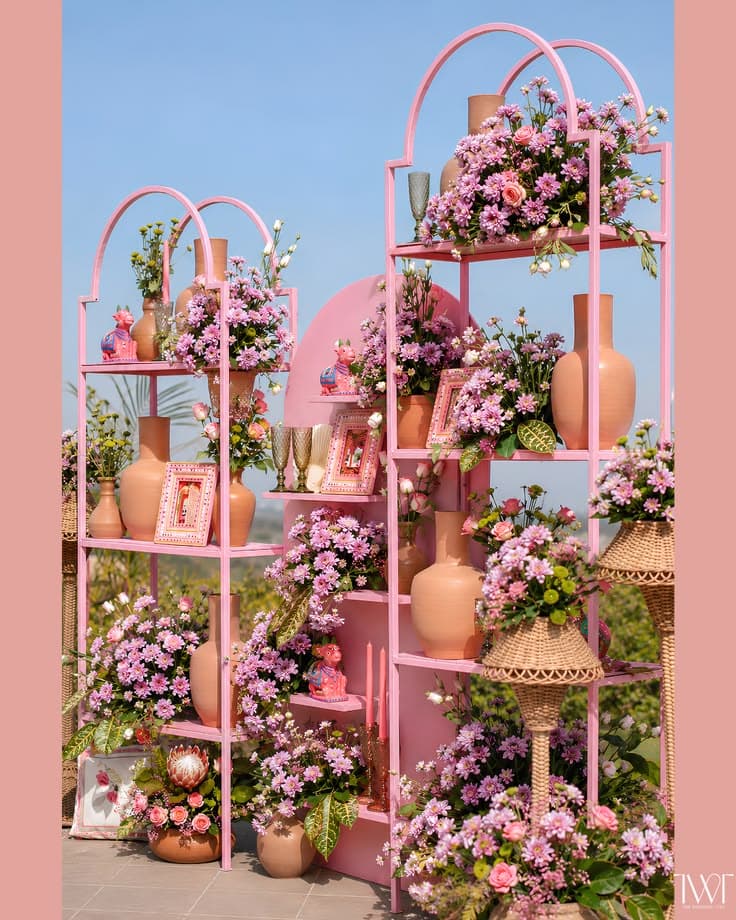 A decorative, freestanding shelving unit constructed from pink-painted metal, featuring a multi-tiered design with elegant arched tops. The unit is styled with numerous floral arrangements in terracotta vases, picture frames, and small decorative objects. — 3D model