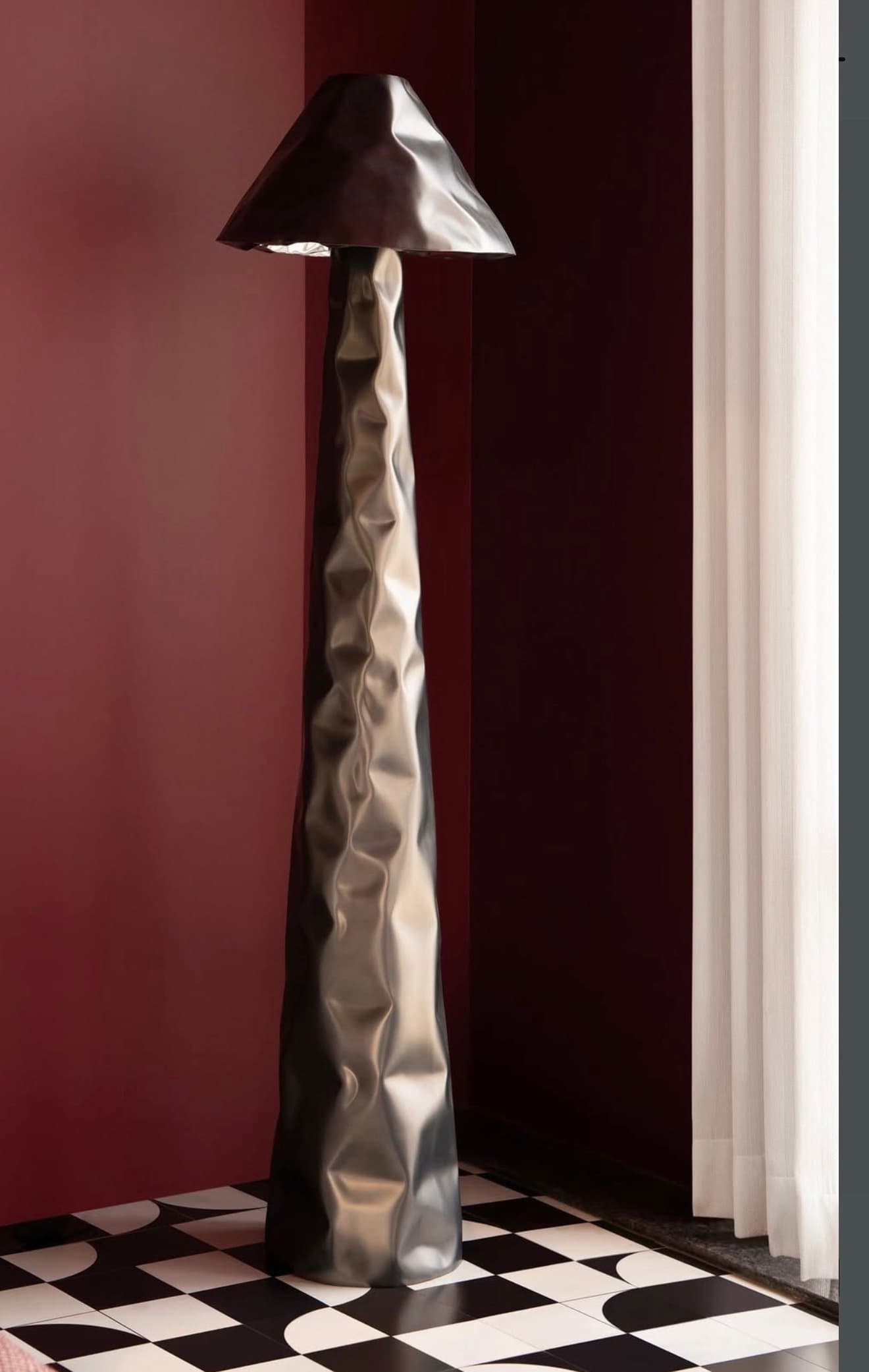 A tall, slender floor lamp crafted from metallic material, featuring a distinctive crumpled or crinkled texture across both its cylindrical base and wide conical shade. The finish appears to be a dark, gunmetal grey, giving it a modern and sculptural aesthetic. The light source is housed within the shade, directing illumination downwards. — 3D model