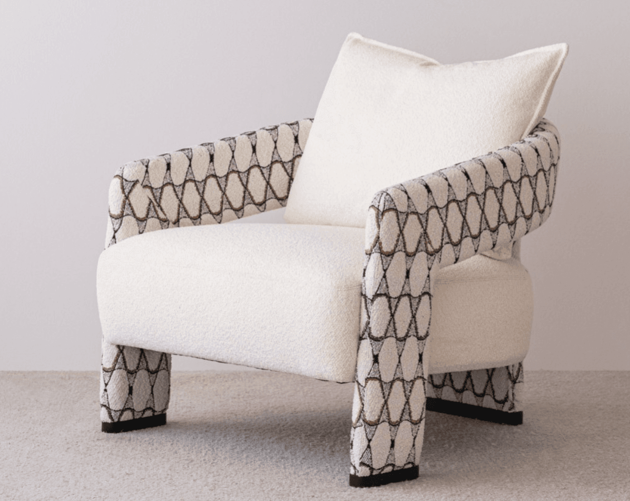 A contemporary upholstered armchair featuring a plush, light-colored bouclé fabric on the seat and back, complemented by patterned armrests and legs with a geometric design in black, white, and a coppery tone. It includes a matching back cushion. — 3D model
