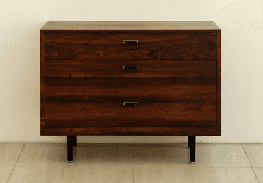 A dark wooden chest of three drawers with slender black legs and minimalist metal handles, set against a light wall. — 3D model