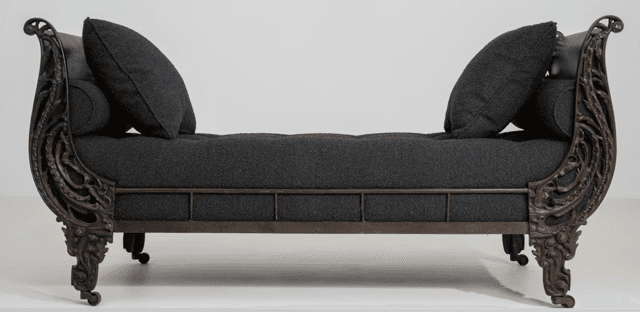 An ornate traditional recamier daybed featuring a dark, possibly bronzed or antiqued metal frame with intricate scrolled and carved details on both raised ends and the legs. The main seating surface is a deep button-tufted cushion upholstered in a dark gray or charcoal fabric. It is accompanied by a matching square throw pillow and a cylindrical bolster pillow. — 3D model