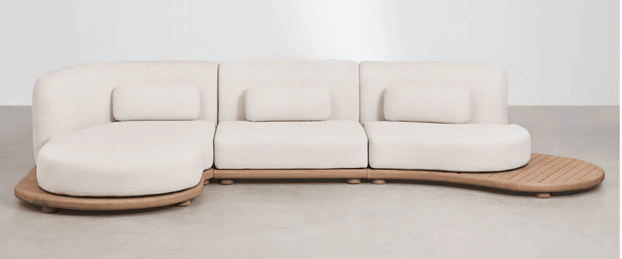 A modular sectional sofa with a curved chaise lounge section and a detached wooden table extension. The sofa features a low profile base and rounded edges. It is upholstered in a light-colored fabric and includes several rectangular throw pillows. — 3D model