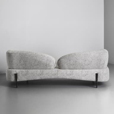 A close-up view of a segment of a modern sofa, featuring rounded, organic lines and textured upholstery. The sofa has a plush appearance, with a low profile and substantial cushioning. A single, distinctly designed metal leg is visible, supporting the rounded front base of the sofa. The leg has a dark, textured finish with a cap that appears to be a different material, possibly metal or wood with a metallic finish. — 3D model