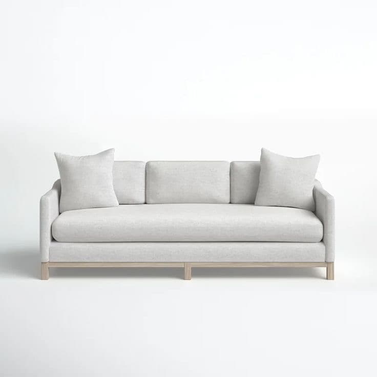 A contemporary three-seater sofa featuring light grey textured upholstery, track arms, and an exposed light-toned wooden base. It includes three loose back cushions and two matching throw pillows. — 3D model