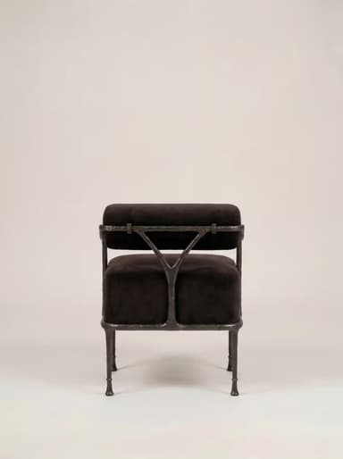 A robust armchair featuring a dark, possibly black or very dark brown, velvet upholstered seat and a cylindrical backrest. The frame is constructed from dark, textured, possibly forged iron or a similar heavy metal, exhibiting a somewhat raw and organic aesthetic. The legs are slender but robust, tapering slightly to small, disc-like feet. The backrest is suspended above the seat, supported by the frame's uprights, with a small V-shaped support element visible at the rear. — 3D model