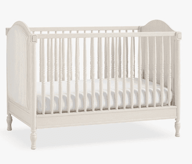 A light-colored, traditional style wooden crib featuring turned spindles and paneled end posts. The finish appears distressed or washed, giving it a soft, aged aesthetic. A white mattress is visible inside. — 3D model