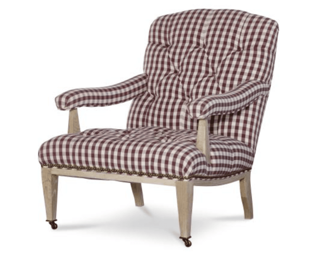 A traditional upholstered armchair featuring a maroon and white gingham check pattern. The backrest and seat are deeply tufted, providing a classic, cushioned appearance. The armrests are also upholstered and gently rolled, supported by exposed light-toned wooden stiles. The chair's frame includes visible wooden legs, with the front legs equipped with brass casters for mobility. Decorative brass nailhead trim outlines the base of the seat where the fabric meets the wooden frame. — 3D model