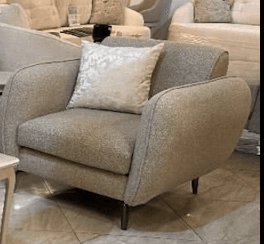 A modern upholstered armchair featuring soft, rounded lines and a light beige or greige textured fabric. The chair includes a matching throw pillow with a subtle patterned design. It rests on four slim, tapered dark metal legs. — 3D model