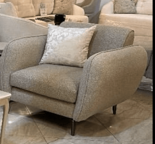 A modern upholstered armchair featuring soft, rounded lines and a light beige or greige textured fabric. The chair includes a matching throw pillow with a subtle patterned design. It rests on four slim, tapered dark metal legs. — 3D model