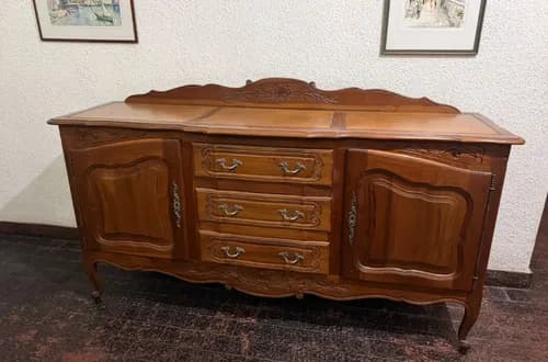 A classic French Provincial style wooden sideboard with a rich, warm brown finish. It features a serpentine front, cabriole legs, and ornate brass hardware. The top edge is scalloped, and intricate floral and scroll carvings adorn the drawer fronts and side cabinet panels. The piece offers three central drawers flanked by two cabinet doors, providing ample storage. — 3D model