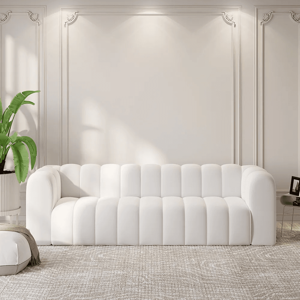 A white, plush sofa with a channel-tufted design across the back and seat cushions. The sofa features rounded armrests and a streamlined silhouette, suggesting a modern or contemporary aesthetic. The lighting highlights the vertical channels, creating a subtle play of light and shadow. — 3D model