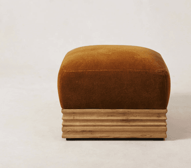 A square-shaped ottoman with a plush, upholstered top and a fluted wooden base. The upholstery is a deep, rich ochre or burnt orange color with a soft texture, possibly velvet. The base is constructed from wood with a natural finish, featuring horizontal, carved grooves that give it a ribbed appearance. The overall design blends soft, tactile materials with a structured, textured base. — 3D model