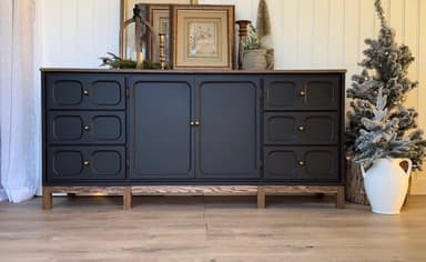 A contemporary, long, and low sideboard cabinet featuring a dark wood top surface contrasting with a black-painted body. The cabinet includes two sections of three drawers each, positioned on either side of a central double-door cabinet. All drawers and doors are adorned with small brass knob pulls. The drawer fronts have a distinctive recessed or carved panel detail. The entire unit is elevated on a simple, dark wood base with short, sturdy legs. — 3D model