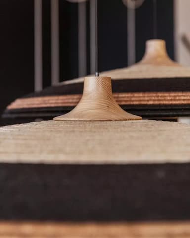 A close-up image of two pendant lights, each featuring a bell-shaped natural wood canopy from which a woven natural fiber shade hangs. The shades are multi-toned with alternating bands of light beige/natural and black woven material, creating a striped pattern. The wooden canopies exhibit a visible grain, and the black electrical cord is visible emerging from the top of the canopy. — 3D model