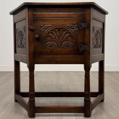 A vintage wooden cabinet with a triangular top. The front features carved decorative elements, a hinged door with a small knob handle, and metal hinges. The cabinet has four legs, two of which are visible, and a stretcher connecting them near the bottom. The wood grain is prominent, and the finish is dark brown. It appears to be a small accent cabinet or a cellarette. — 3D model