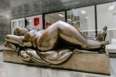 A large bronze sculpture depicting a reclining female figure, situated outdoors on a substantial concrete plinth in front of a modern glass building. The figure has soft, rounded forms and is posed with her head resting on a raised form resembling a pillow, with her body gently draped. — 3D model