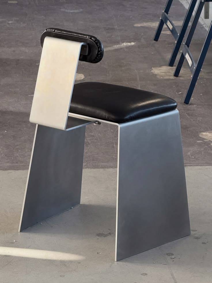 An armless chair with a minimalist design, featuring a bent metal frame in a matte silver finish and a black faux leather upholstered seat and a small backrest. — 3D model