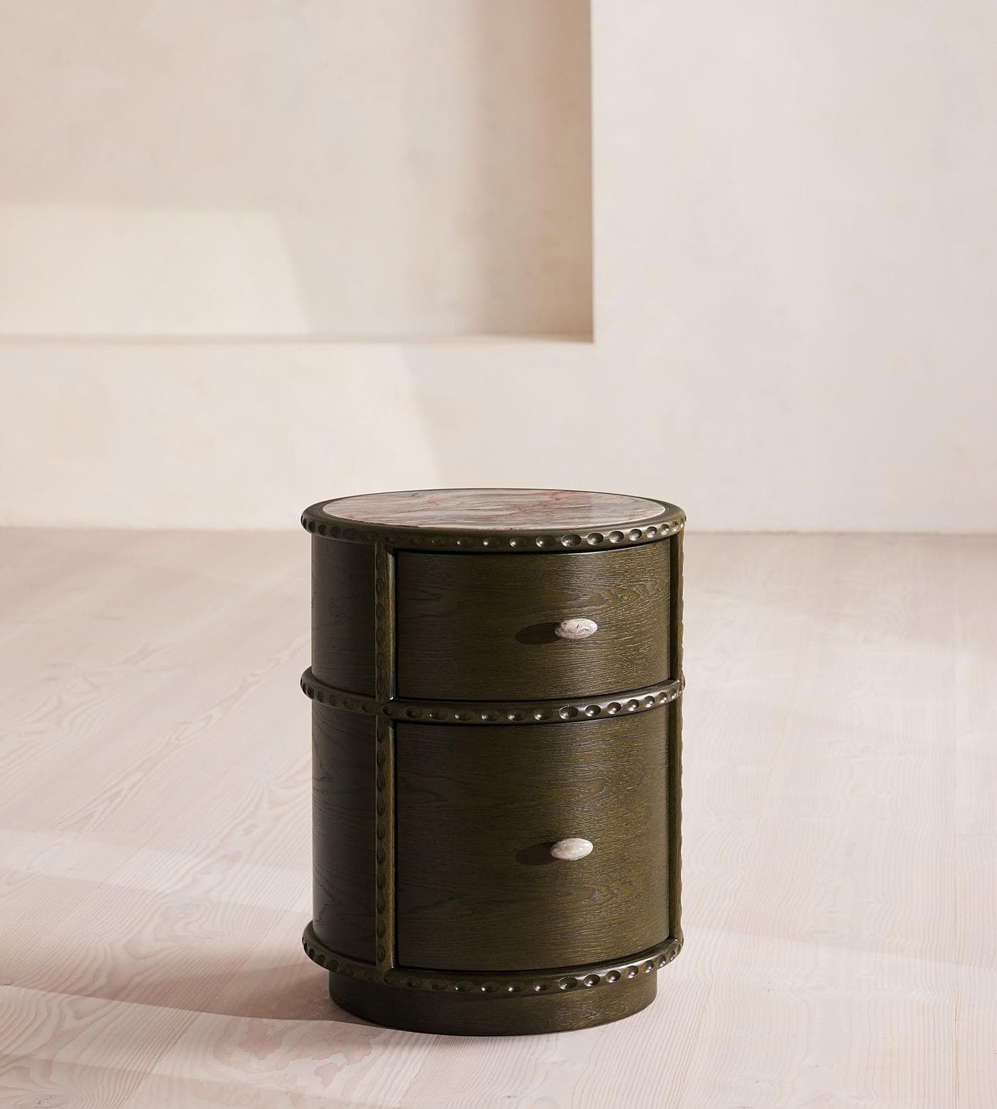 A cylindrical nightstand with a marble top and two drawers. The body is constructed from dark stained wood with visible grain. Decorative metal accents, resembling rivets or studs, adorn the top and bottom edges of the cylinder and separate the two drawers. Each drawer has a single oval knob that appears to be made of a light-colored stone. — 3D model
