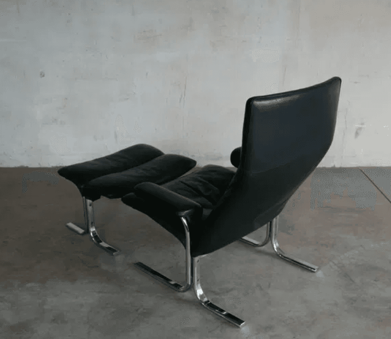 A black leather armchair with a matching ottoman. The chair features a high back and a curved, cantilevered chrome base. The ottoman also has a curved chrome base and is upholstered in matching black leather with a segmented cushion design. — 3D model