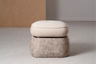 A two-tiered ottoman with a rounded, tufted top cushion and a patterned base. The top cushion is upholstered in a smooth, light beige fabric and features a single button tuft in the center, outlined by stitched seams. A contrasting piping trim defines the edge of the top cushion. The base is wider than the top and is upholstered in a textured fabric with a repeating abstract pattern in shades of beige and white, resembling arcs and linear shapes. The ottoman is situated on a textured, light-colored carpet. — 3D model