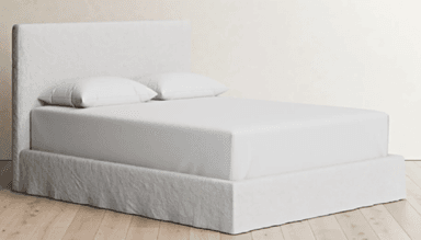 A modern upholstered platform bed featuring a simple, rectangular headboard and a fabric-wrapped bed frame. The entire structure, including the headboard and base, is covered in a light-colored, possibly linen or cotton, fabric with a slightly textured finish. The bed includes a mattress and two pillows, all covered in plain white sheets, resting within the upholstered frame. The design is minimalist and clean-lined, with the fabric covering extending to the floor around the base. — 3D model