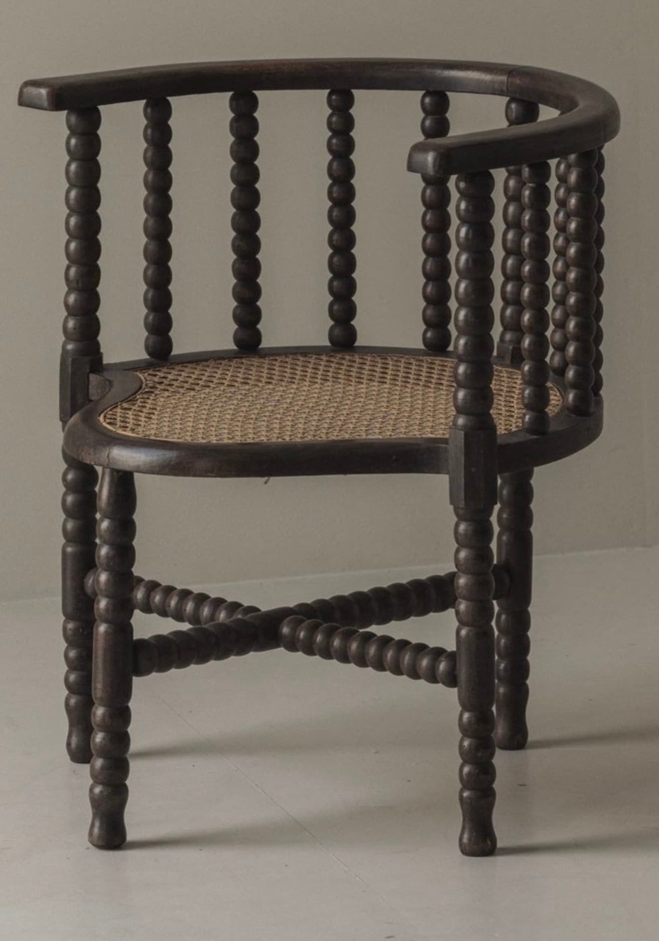 A dark stained wooden barrel armchair featuring a bobbin-turned frame and a woven cane seat. The back and armrests form a continuous curved structure, supported by vertical bobbin-turned spindles. The four bobbin-turned legs are connected by bobbin-turned stretchers forming an X-shape beneath the seat. The seat itself is crafted from woven cane with a slightly curved front. — 3D model