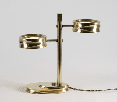 A modern two-light table lamp constructed from brass, featuring a central vertical stem with two horizontally extending arms, each supporting a cylindrical light fixture with a distinctive fluted or reeded exterior texture. The base is an oval, layered design with similar fluted detailing. The lamp is illuminated, showcasing integrated LED light sources, and includes a braided fabric power cord. — 3D model