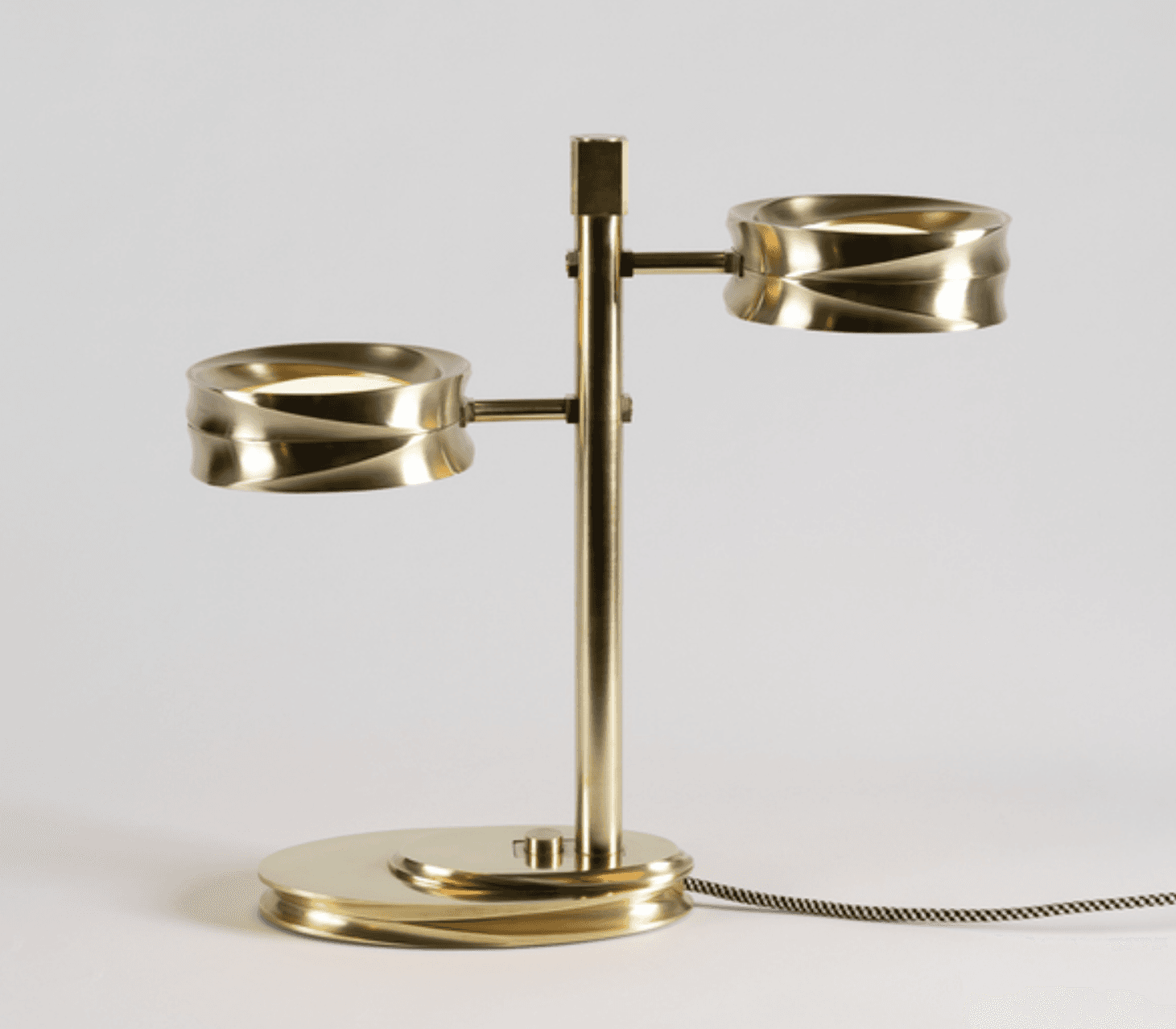 A modern two-light table lamp constructed from brass, featuring a central vertical stem with two horizontally extending arms, each supporting a cylindrical light fixture with a distinctive fluted or reeded exterior texture. The base is an oval, layered design with similar fluted detailing. The lamp is illuminated, showcasing integrated LED light sources, and includes a braided fabric power cord. — 3D model