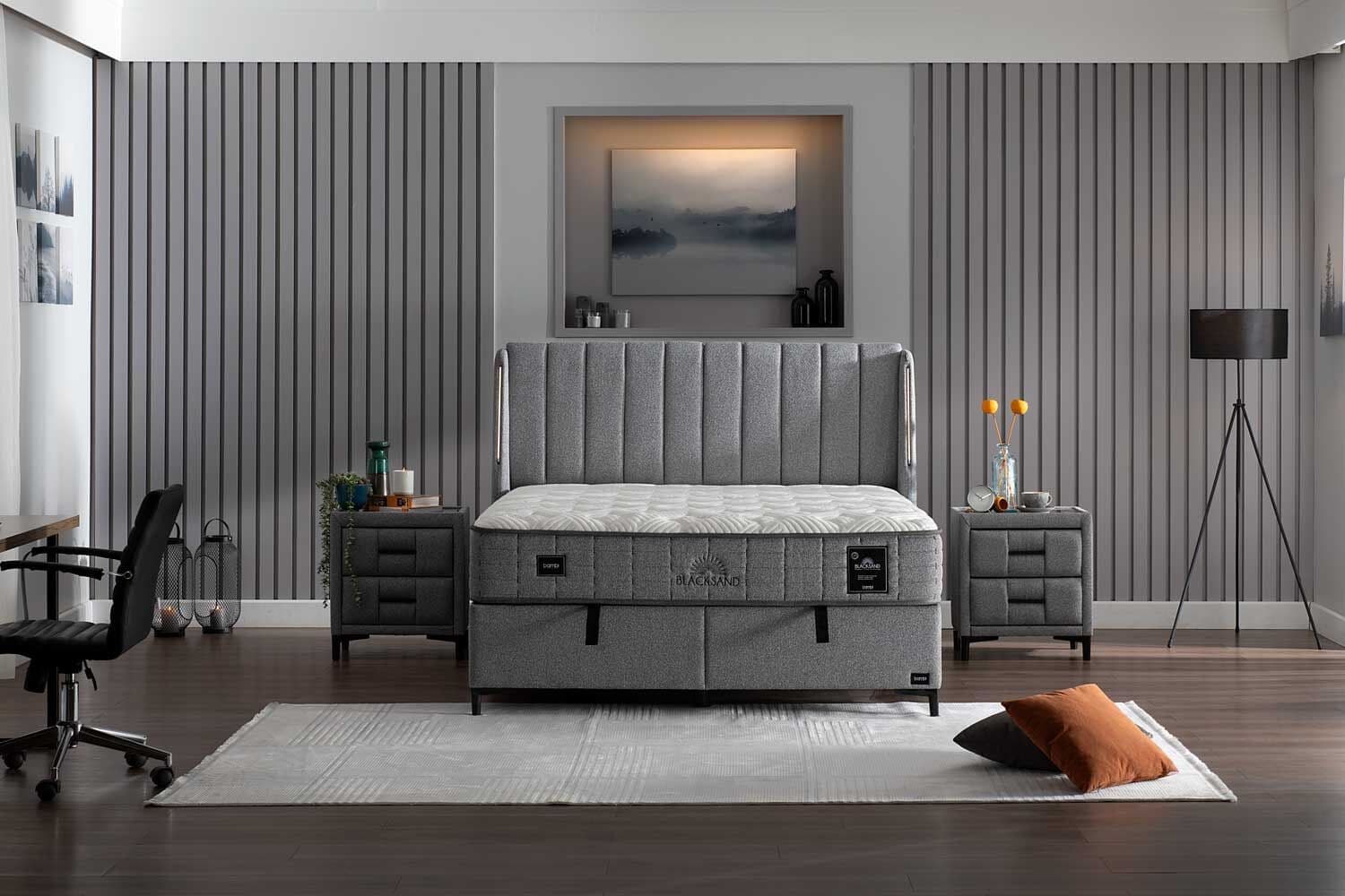 A modern upholstered bed frame with a tufted headboard and a mattress. The bed frame is upholstered in a textured gray fabric and features two integrated drawers at the foot of the bed. The mattress is white with a patterned quilted top and a gray border. The headboard has vertical channel tufting. The bed is supported by slender black legs. — 3D model