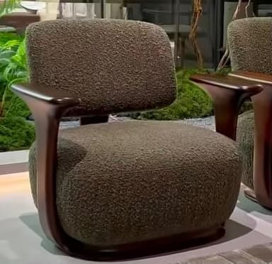 A contemporary armchair featuring a plush, textured olive-green upholstery, supported by a distinct dark wood frame that forms the armrests and wraps around the base. The design is characterized by its rounded, ergonomic forms and clean lines. — 3D model