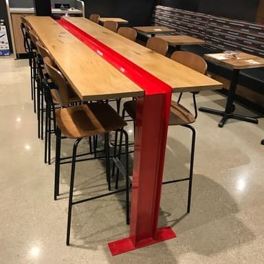 A long bench featuring a light-colored wooden top and a vibrant red metal frame. The wooden top is composed of two planks separated by a central red metal channel. The frame consists of two sturdy, upright red metal supports that resemble I-beams, each with a flat base plate designed for anchoring to the ground. — 3D model