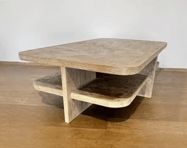 A rectangular coffee table featuring softly rounded corners and a textured, light beige or gray finish that mimics the appearance of concrete or plaster. It includes a recessed lower shelf for additional storage or display, supported by a central cross-shaped base. — 3D model