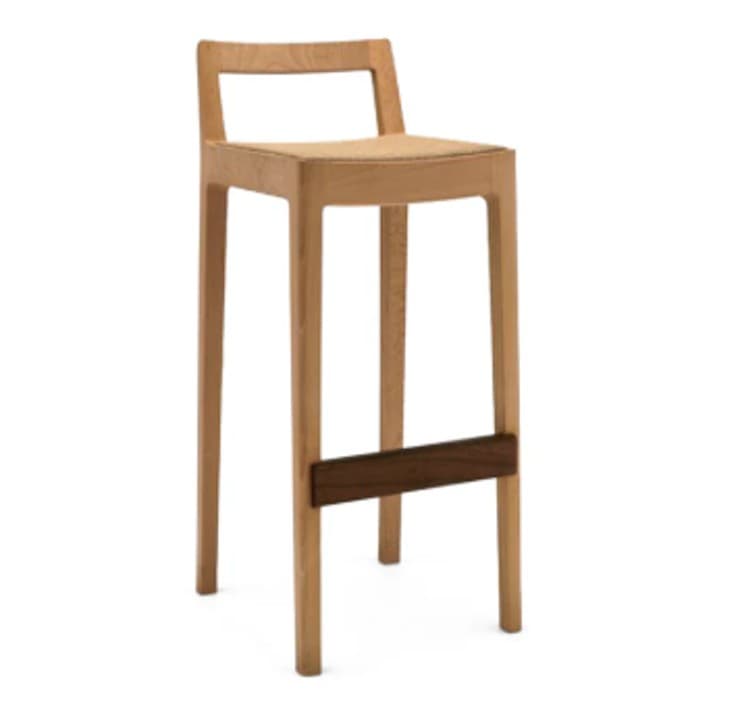 A modern wooden bar stool featuring a light-toned wood frame, a square open backrest, and a woven rattan seat. A contrasting dark wood footrest is positioned between the front legs. — 3D model