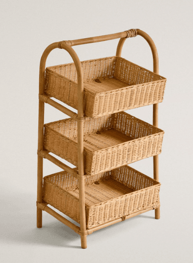A three-tiered storage unit constructed primarily from natural rattan and woven wicker. The frame is made of curved and straight rattan poles, creating three levels, each holding a rectangular woven wicker basket. The top of the unit features an integrated arched handle, suggesting portability. The overall aesthetic is natural and bohemian, with a light brown, natural finish. — 3D model