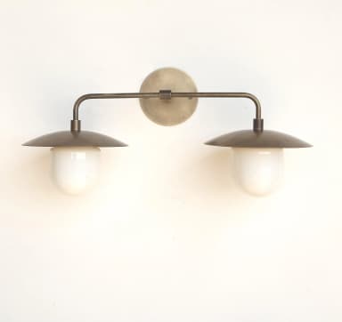 A wall-mounted lighting fixture featuring two light sources, constructed from aged brass or bronze metal, with each light housed under a conical metal shade and diffusing light through an opaque, white, bell-shaped glass component. — 3D model
