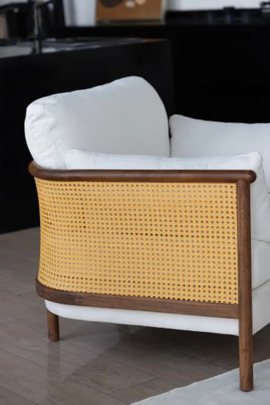 A modern lounge chair featuring a curved dark wood frame with cane webbing on the backrest, complemented by plush, off-white fabric cushions. The chair is designed for comfort and adds a relaxed yet elegant aesthetic to an interior space. — 3D model