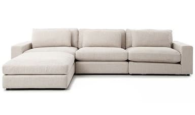 A modern sectional sofa featuring a modular design with a left-facing chaise lounge. The sofa is upholstered in a light beige or off-white woven fabric, presenting a clean and minimalist aesthetic. It comprises several connected units, including a chaise, two armless seats, and one seat with a right armrest. Each seating segment features thick, boxy seat cushions and loose back cushions, along with smaller throw pillows. The armrests are broad and flat, contributing to the sofa's contemporary block-like silhouette. The overall design emphasizes comfort and clean lines, suitable for a relaxed living space. — 3D model