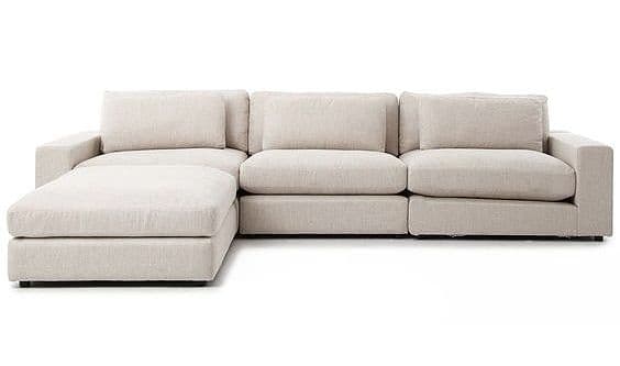A modern sectional sofa featuring a modular design with a left-facing chaise lounge. The sofa is upholstered in a light beige or off-white woven fabric, presenting a clean and minimalist aesthetic. It comprises several connected units, including a chaise, two armless seats, and one seat with a right armrest. Each seating segment features thick, boxy seat cushions and loose back cushions, along with smaller throw pillows. The armrests are broad and flat, contributing to the sofa's contemporary block-like silhouette. The overall design emphasizes comfort and clean lines, suitable for a relaxed living space. — 3D model