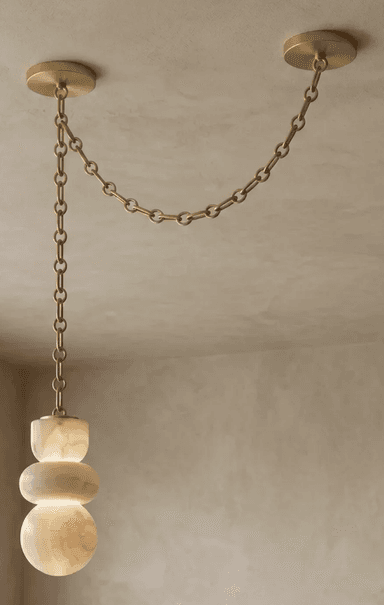 A contemporary pendant light featuring a sculptural shade crafted from translucent alabaster, suspended by a black metal chain system. The shade is composed of three stacked, organic, rounded forms, illuminated from within to highlight the natural veining and luminous quality of the stone. The suspension system consists of a long chain dropping from one ceiling mount, connecting to the light fixture, and a shorter chain extending horizontally to a second ceiling mount, creating a distinctive draped effect. — 3D model