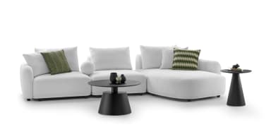 An L-shaped sectional sofa in a white, textured fabric, consisting of multiple modular components, including a chaise lounge end. The sofa features plush, rounded cushions and backrests, accompanied by several matching white throw pillows and three green patterned accent pillows. The design is low-profile and contemporary. — 3D model