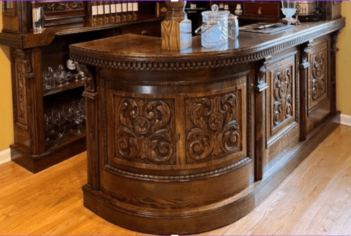An ornate, dark brown wooden bar counter featuring a curved design and intricate relief carvings. The counter is constructed from dark-stained wood, possibly mahogany or a similar rich tone, and includes decorative fluted columns and detailed moldings along its base and top edges. The front panels are adorned with floral and scrollwork carvings, lending it a traditional or classic aesthetic. The top surface is smooth and polished, designed for serving. The overall appearance suggests a high-quality, handcrafted piece. — 3D model