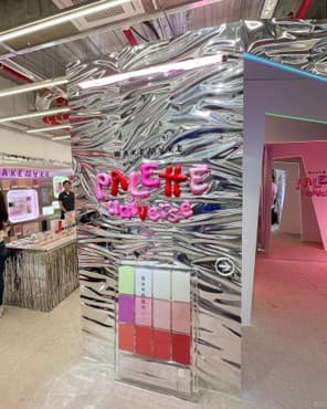 A modern, upright cosmetic retail display stand featuring a textured, silver-grey panel. The panel has a rippled or wavy surface, resembling concrete or a distressed metallic finish. A clear acrylic holder is attached to the lower section, designed to showcase multiple cosmetic palettes. Above the palettes, the brand name 'WAKEMYKE' and 'PALETTE Universe' are rendered in three-dimensional, bubbly pink and red lettering. A thin, horizontal light strip is integrated into the top edge of the main panel, providing illumination. The overall aesthetic is clean and contemporary, suitable for product presentation in a retail environment. — 3D model