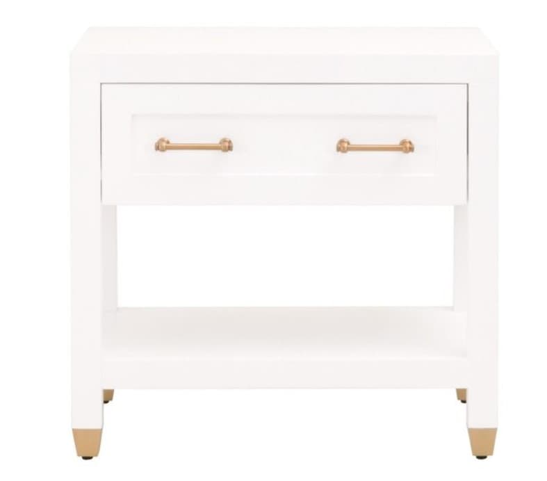 A white nightstand with two drawers and an open shelf below. The drawers have brass-colored handles and the piece rests on four legs with brass-colored caps. The top edge of the nightstand has rounded corners. The surface of the nightstand appears to have a subtle texture. — 3D model