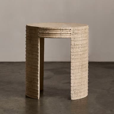 A cylindrical end table with a textured, stone-like surface. The table has a rounded top and a single, curved leg or base element visible. The side of the table exhibits a series of horizontal grooves or ridges, suggesting a layered construction or decorative carving. The material appears porous and natural, consistent with travertine or a similar stone composite. The color is a light, earthy beige. — 3D model